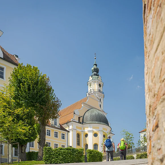 Zwei Personen mit Rucksäcken gehen auf eine gelbe Kirche mit Turm zu, umgeben von Bäumen.