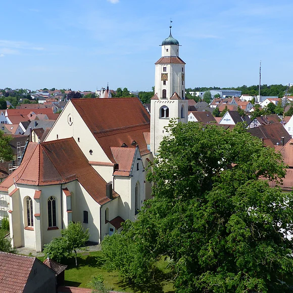 Luftbild der Stadtpfarrkirche mit Turm in einer Stadtlandschaft, umgeben von Häusern mit roten Dächern und Bäumen. Im Hintergrund blauer Himmel.