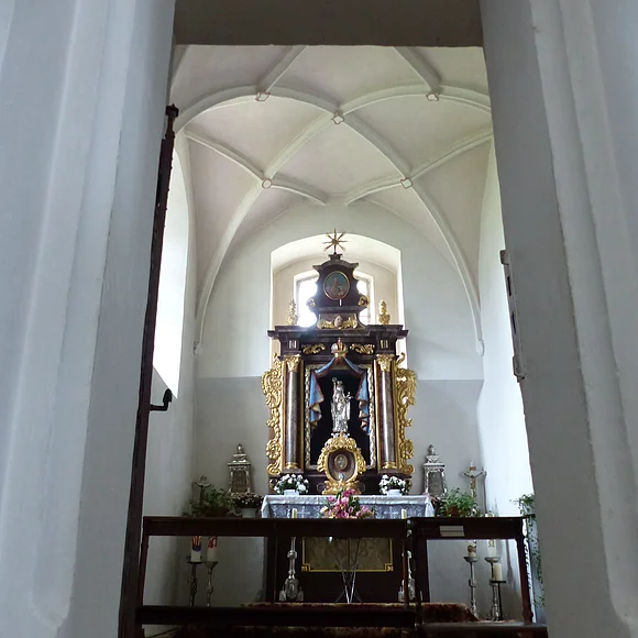 Ein Blick durch eine Tür zu einem Kirchenaltar in der Walburgakapelle in Monheim, mit goldenen Verzierungen und einer Statue, der heiligen Walburga. Der Kirchenaltar ist umgeben von Kerzen und Blumen, in einem gewölbten Raum mit einer verzierten Decke.