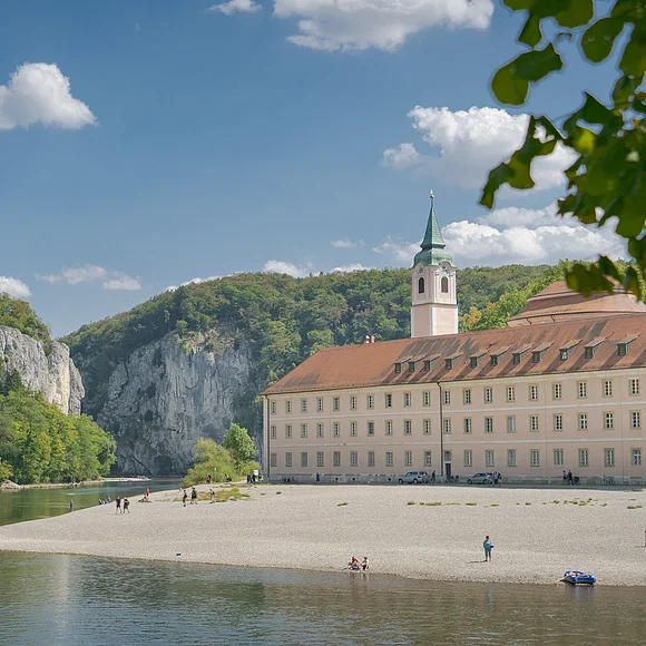 Kloster Weltenburg am Donaudurchbruch Flussufer mit Kiesstrand, altes Gebäude mit Turm und bewaldete Felsen unter blauem Himmel mit Wolken