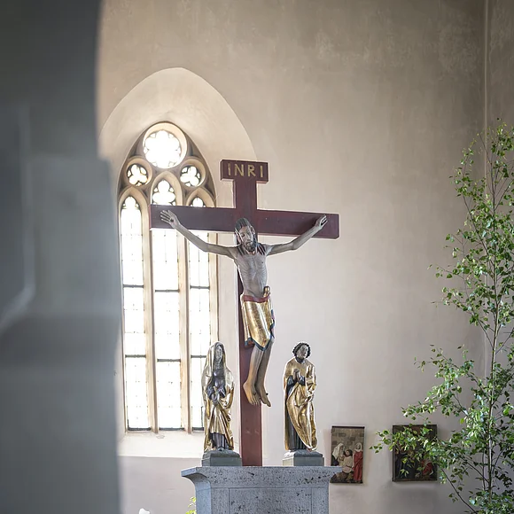 Kruzifix in einer Kirche, flankiert von zwei Figuren. Im Hintergrund ein gotisches Fenster.
