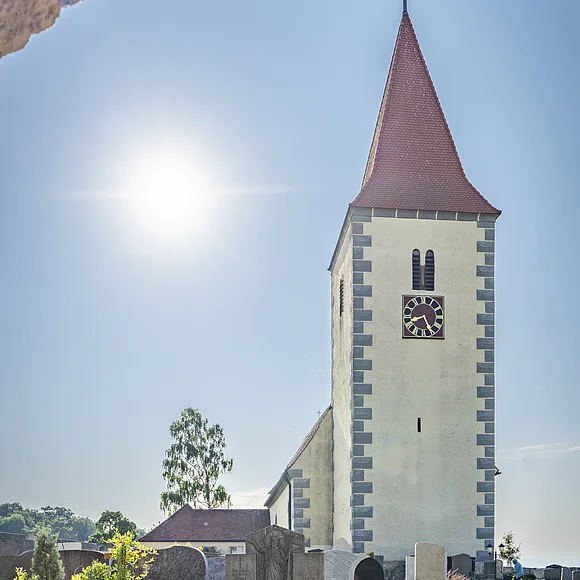Kirchturm mit rotem Dach und Uhr, umgeben von einem Friedhof, unter klarem Himmel mit Sonne.