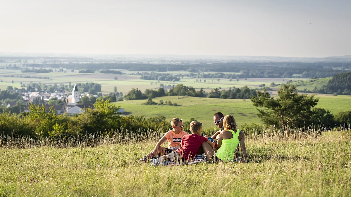 Die Monheimer Alb entdecken - mit herrlichem Ausblick vom Mähhorn in Huisheim Vier Personen sitzen auf einer Wiese mit Blick auf ein Dorf und eine weite Landschaft im Hintergrund.