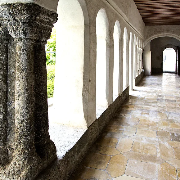 Blick in einen langen Kreuzgang mit Säulen und Rundbögen links im Bild, dahinter grüne Büsche. Rechts im Bild weiße Wand mit einer Ausfräsung in der Wand und Steinplatten mit Text angebracht. Entlang des Gangs alter Bodenbelag aus hellen alten Steinen.