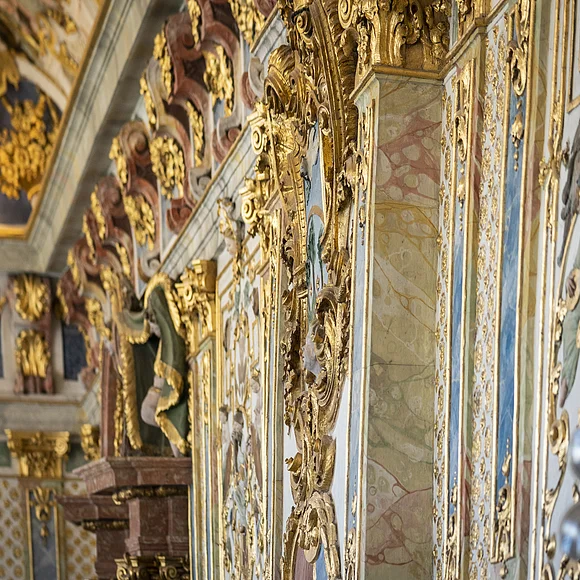 Detailaufnahme einer Wand mit barocker goldener Stuckverzierung und bemalten Figuren in einem historischen Gebäude.