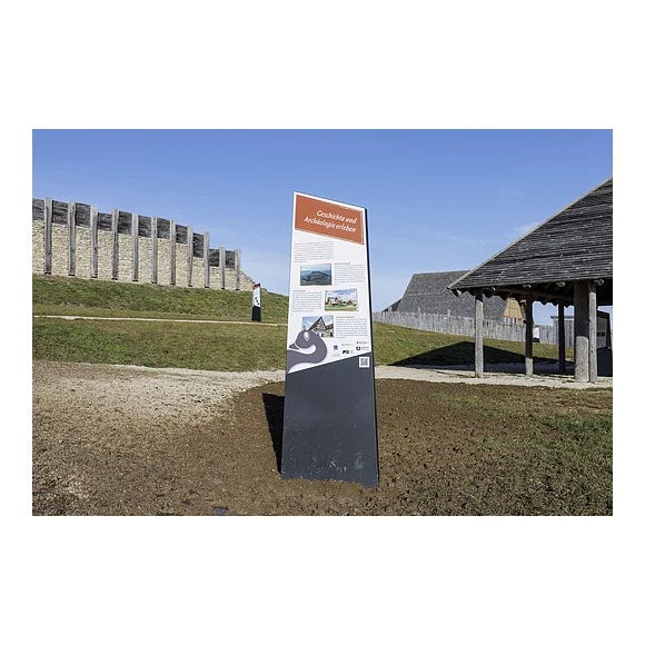 Informationstafel mit dem Titel „Geschichte und Archäologie erleben“ vor einer historischen Holz- und Steinmauer bei klarem Himmel.