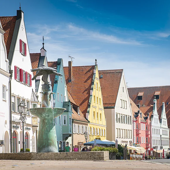 Reichsstraße Donauwörth Straße mit bunten Giebelhäusern und einem Brunnen im Vordergrund. Klarer Himmel im Hintergrund.