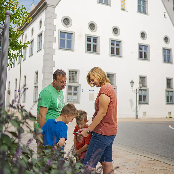 Lauschtour Punkt am Fuggerhaus Eine Familie steht auf einer gepflasterten Straße vor einem historischen Gebäude mit Giebeldach.
