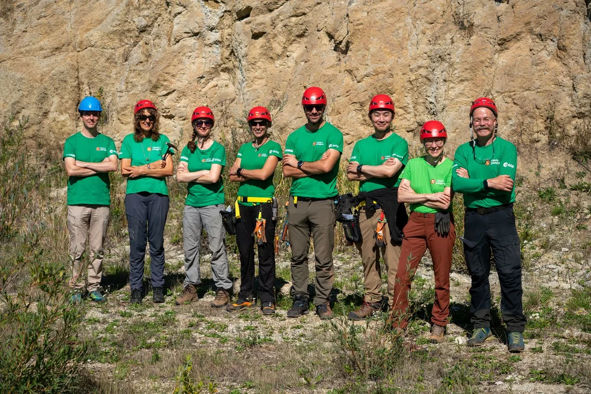 Auf dem Bild sind acht Menschen mit Helmen und grünen T-Shirts zu sehen, die ihre Arme verschränkt haben. Sie stehen im Gras vor einem großen braunen Felsen.